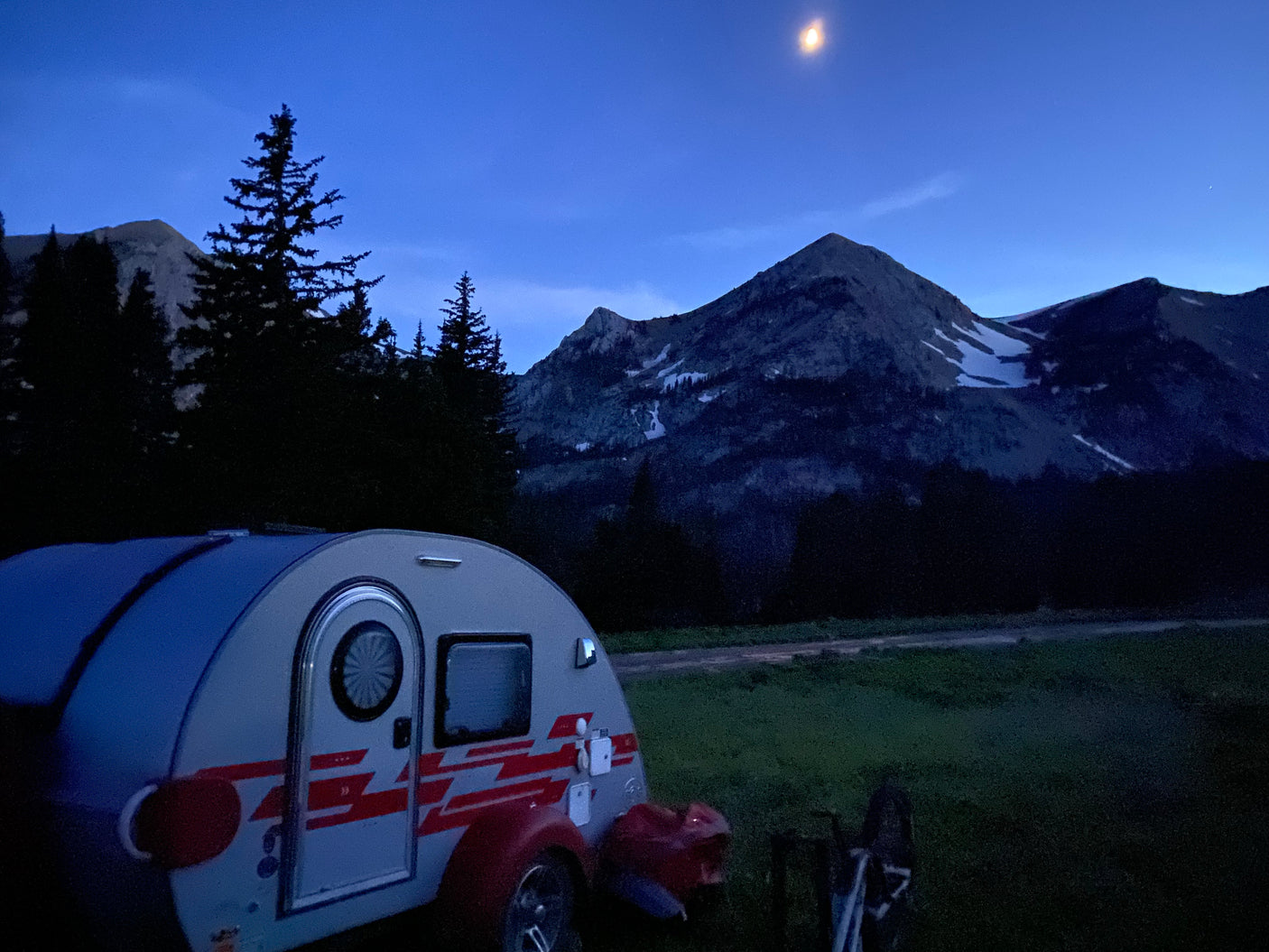Nighttime view: A teardrop RV rental parked amidst towering mountains and dark pine trees under a bright moon.