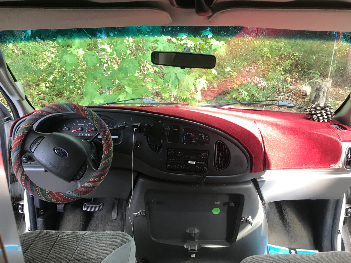Inside an RV rental van: driver's perspective. Features a red dash cover, custom fabric steering wheel, and a lush forest view outside.