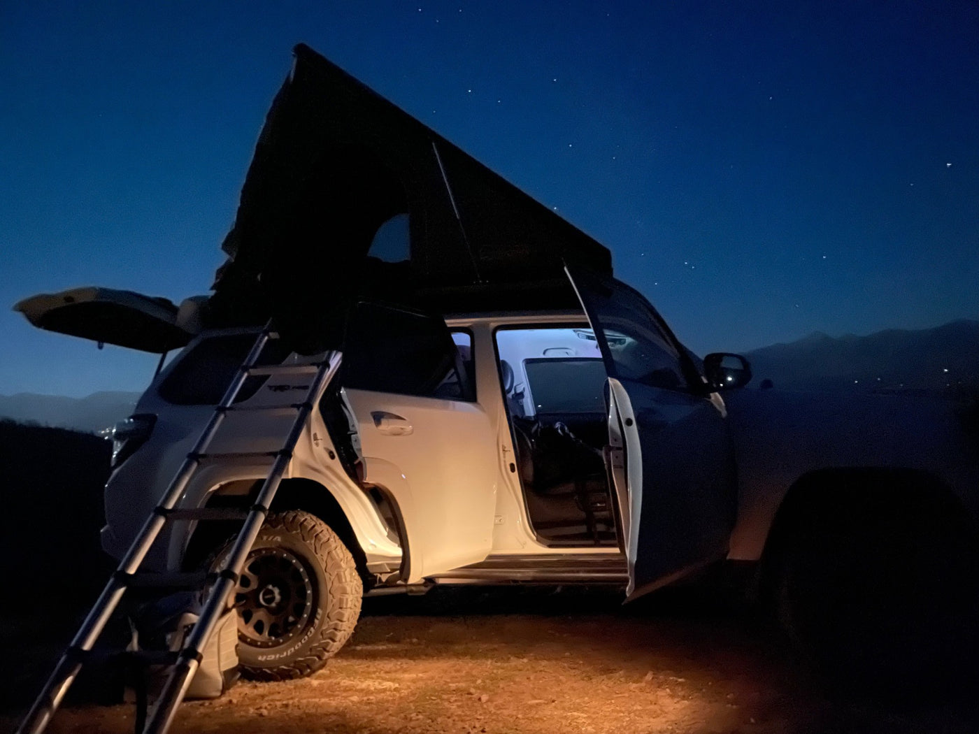 White SUV with rooftop tent & ladder, an ideal RV rental for car camping under a starry night sky. Illuminated interior. — photo 8