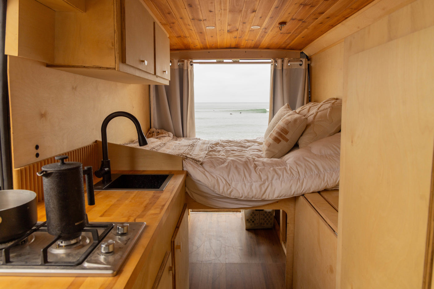 Experience van life in this cozy RV rental: kitchen, bed, and stunning ocean views out the back window.