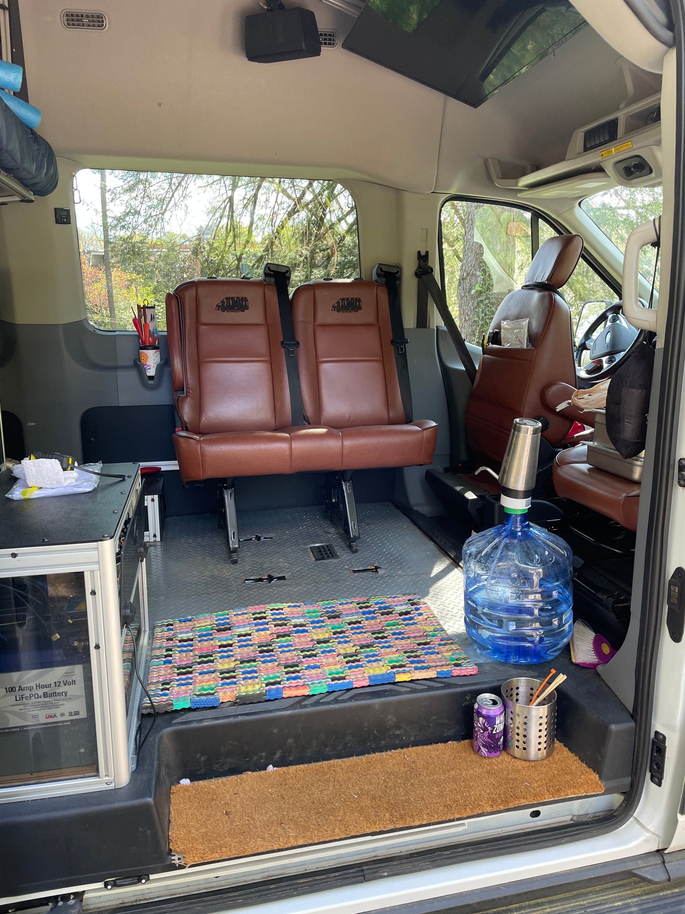 Spacious RV rental interior: brown leather seats, utility station with battery, water dispenser, and colorful floor mats.