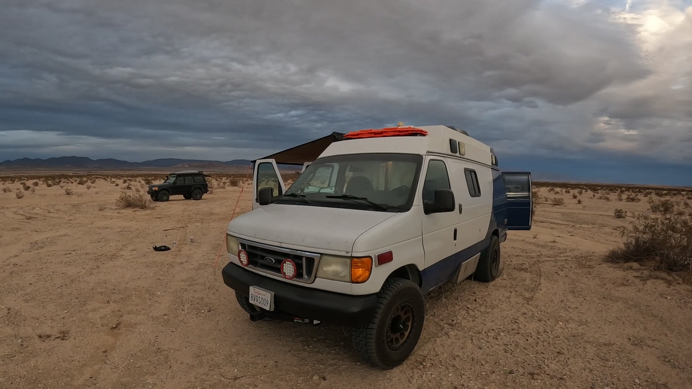 White off-road RV rental van with awning & a second SUV parked in a vast, sandy desert under cloudy skies. Rugged adventures.