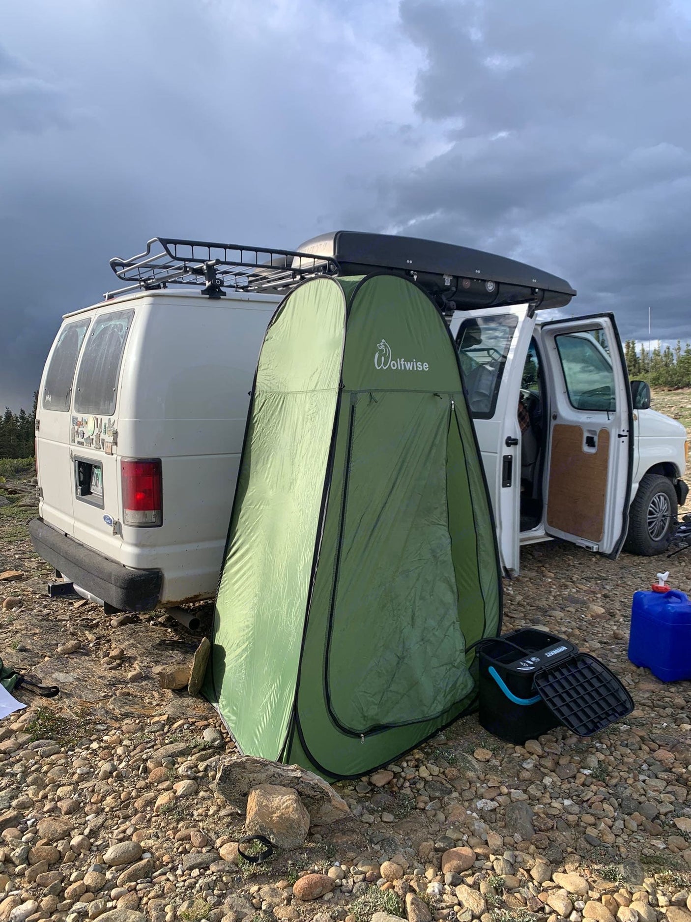 An RV rental van parked on rocky terrain with a privacy tent, portable toilet, and roof storage for remote camping.