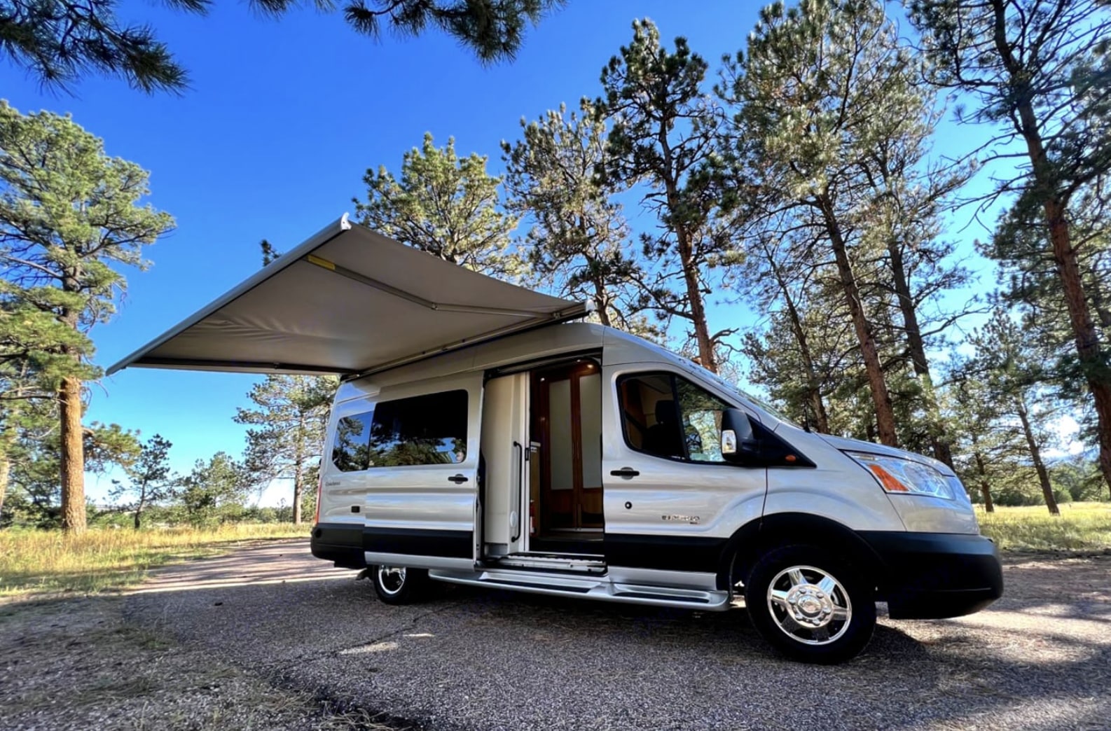 A silver RV van with its door open & awning extended is parked in a sunny pine forest. Your perfect rv rental adventure awaits!