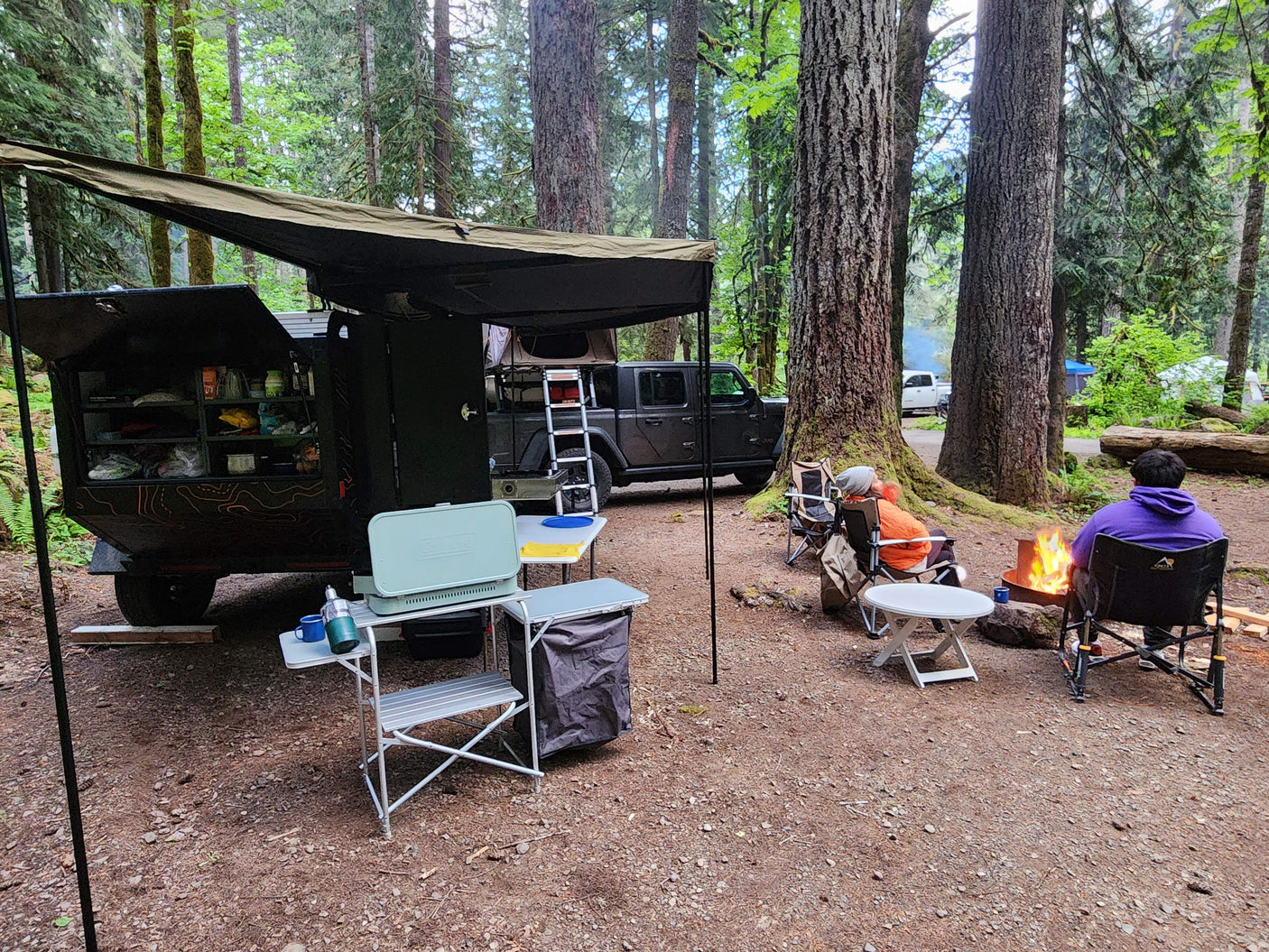 Overlanding setup: dark camper trailer & Jeep. People by a campfire in a forest. Ideal RV rental for outdoor living!