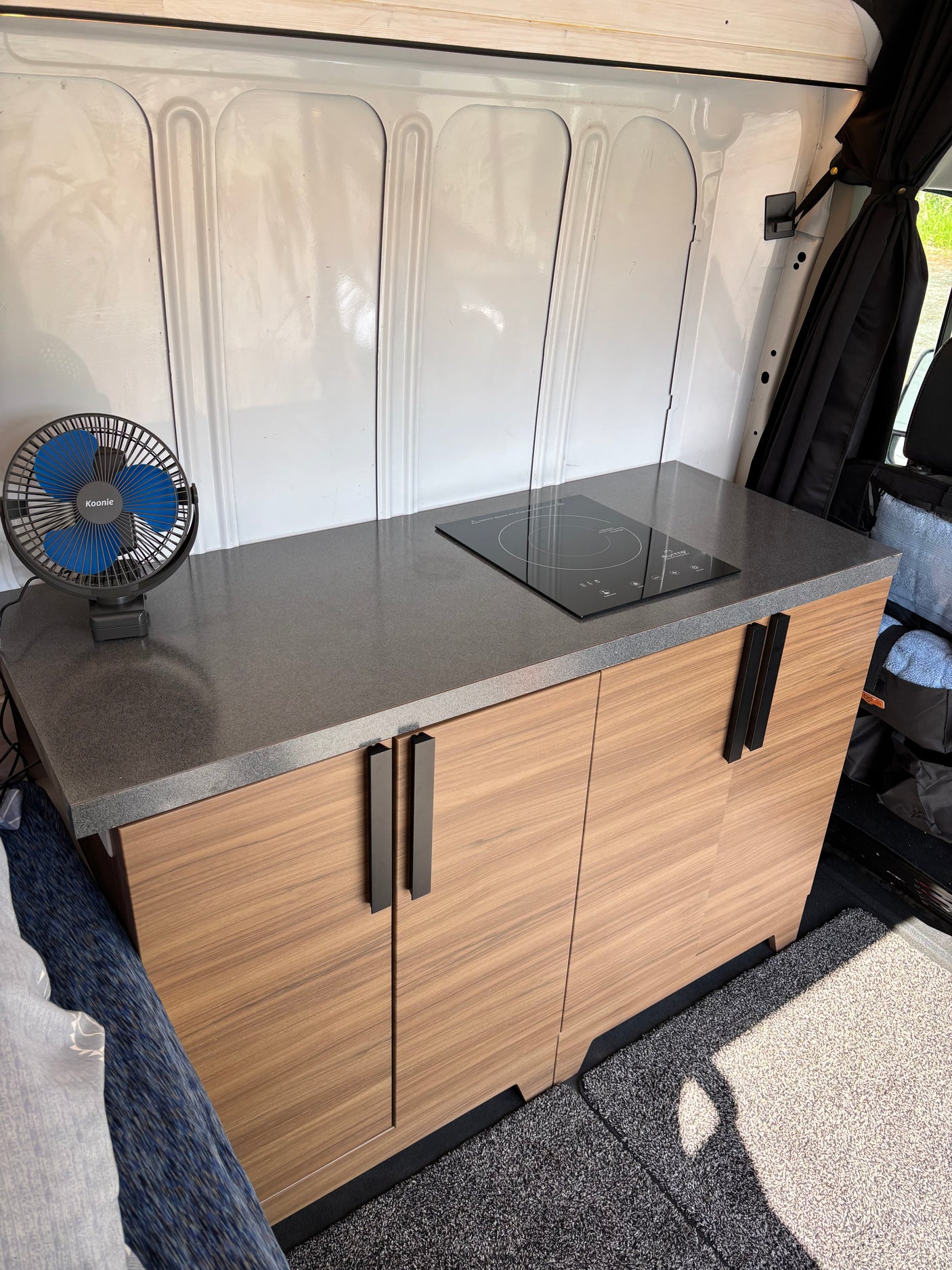 Compact RV rental kitchenette: sleek counter, induction cooktop, and modern wood cabinets in a camper van interior.