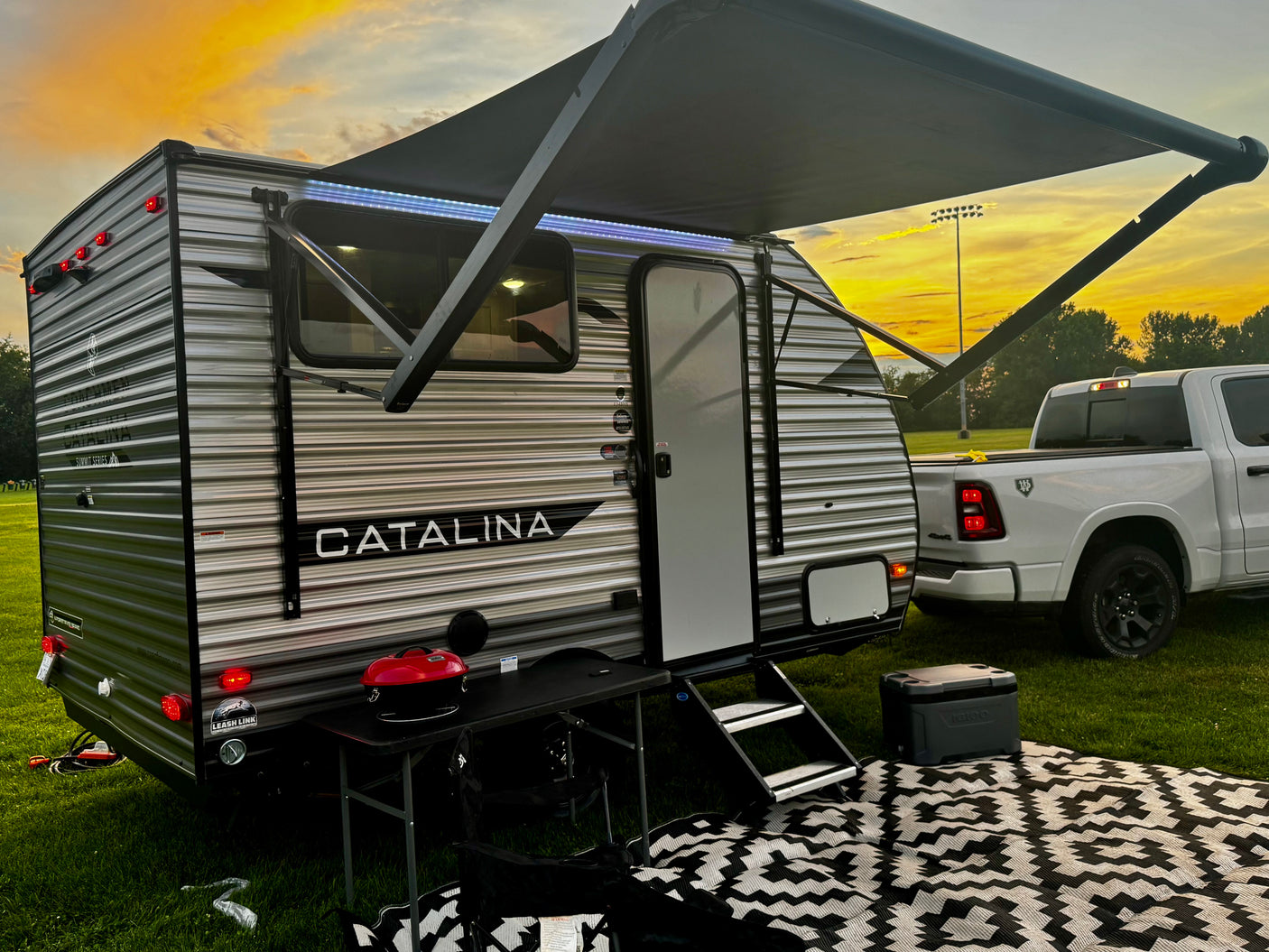 This RV rental, a Catalina travel trailer, is set up in a grassy field with an awning, table, grill, and cooler under a sunset sky.
