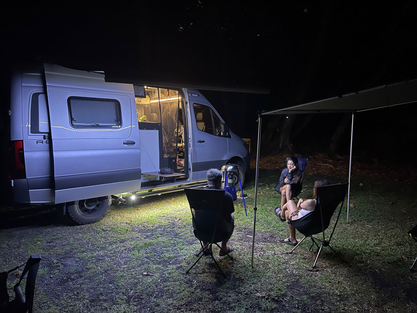 RV rental camper van brightly lit at night. Friends relax in chairs under the awning, enjoying a cozy outdoor camp evening.