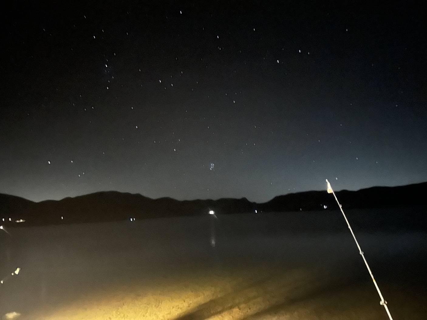 Peaceful night camping with an RV rental. Starlit sky above mountains & water, a fishing rod waits. Perfect for stargazing.