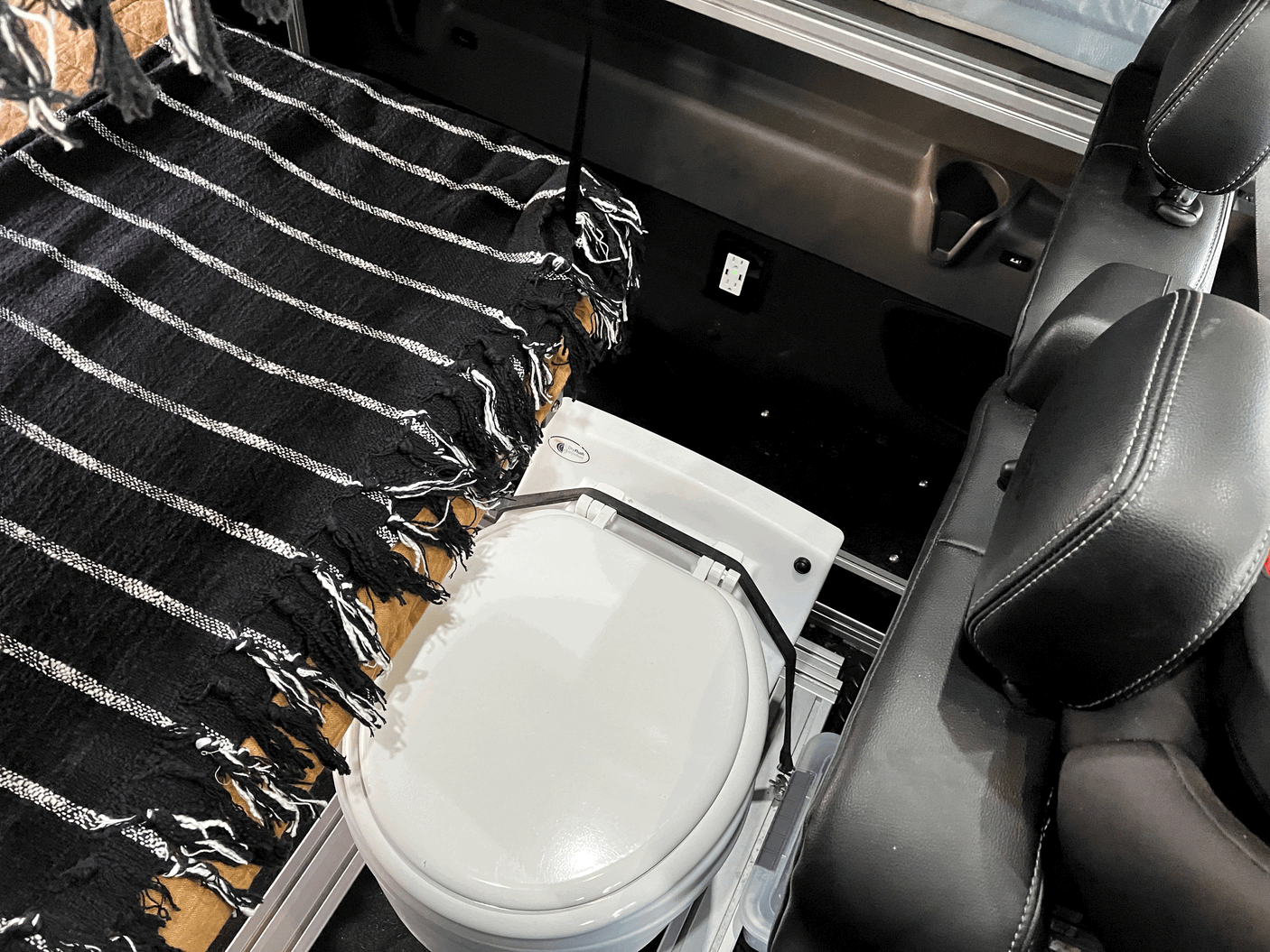Inside an RV rental: view of the portable toilet securely placed next to a black and white blanket and vehicle seats.