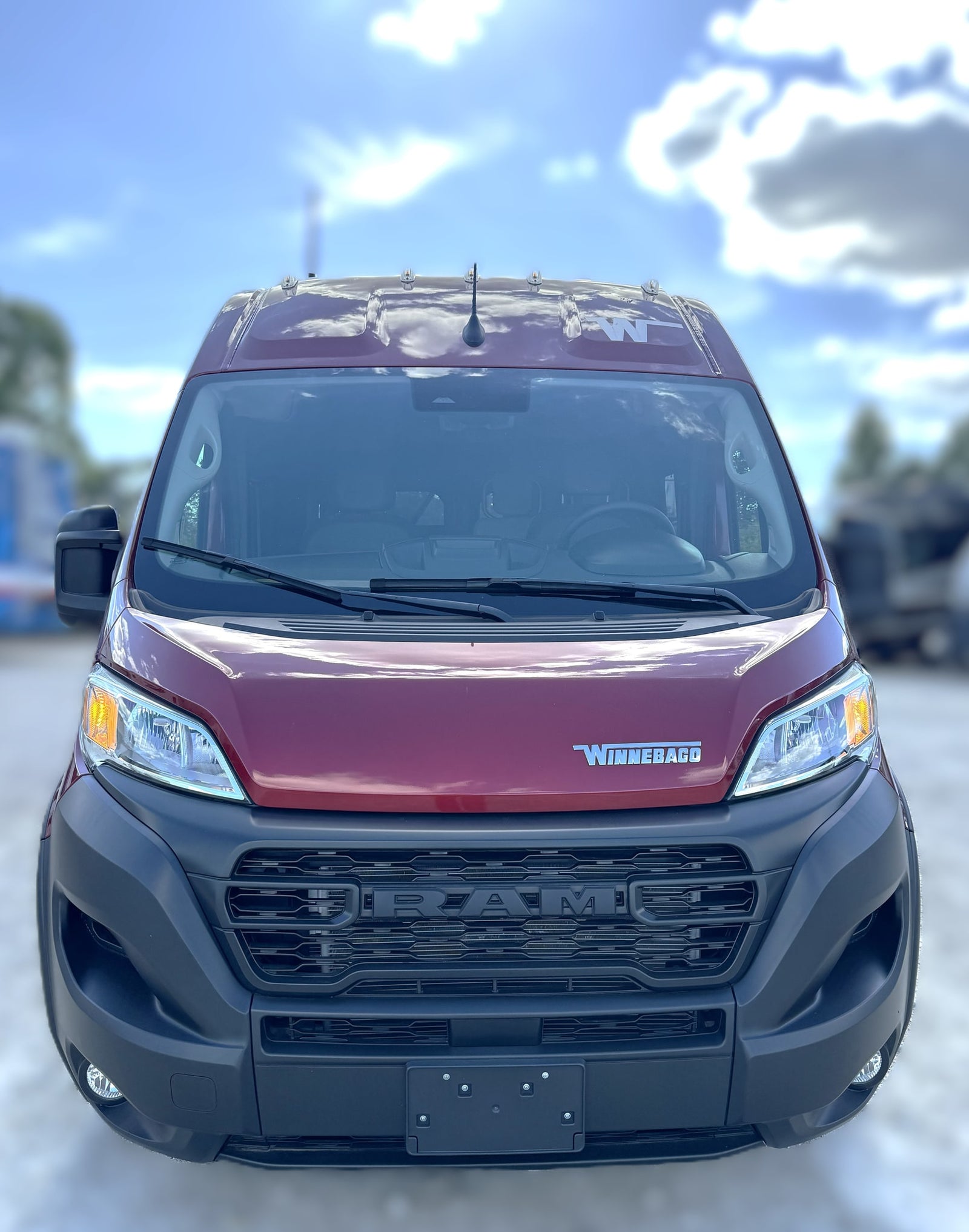 Front view of a red Winnebago RV rental van, based on a Ram ProMaster chassis, parked outdoors under a blue sky.