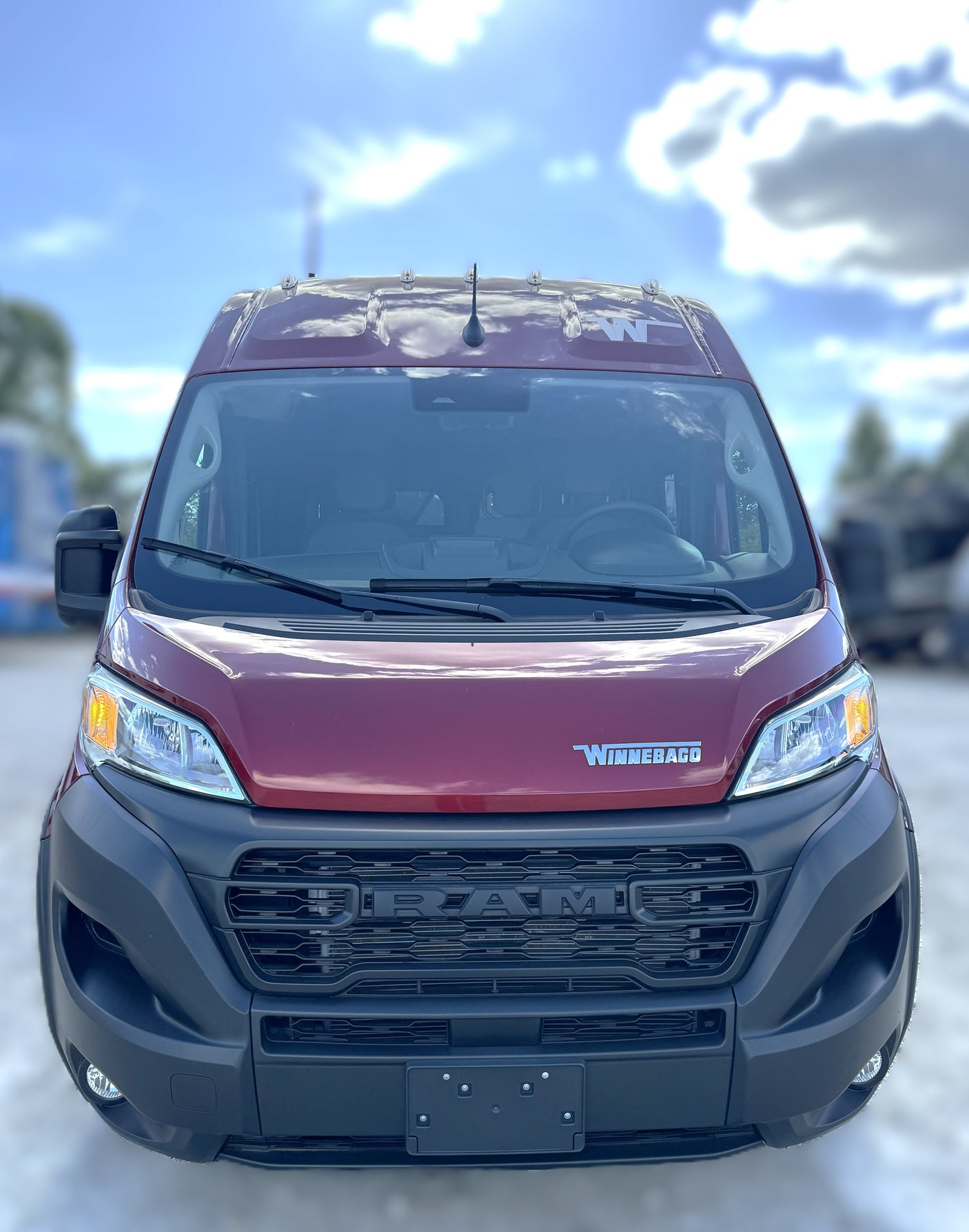 Front view of a red Winnebago RV rental van, based on a Ram ProMaster chassis, parked outdoors under a blue sky.