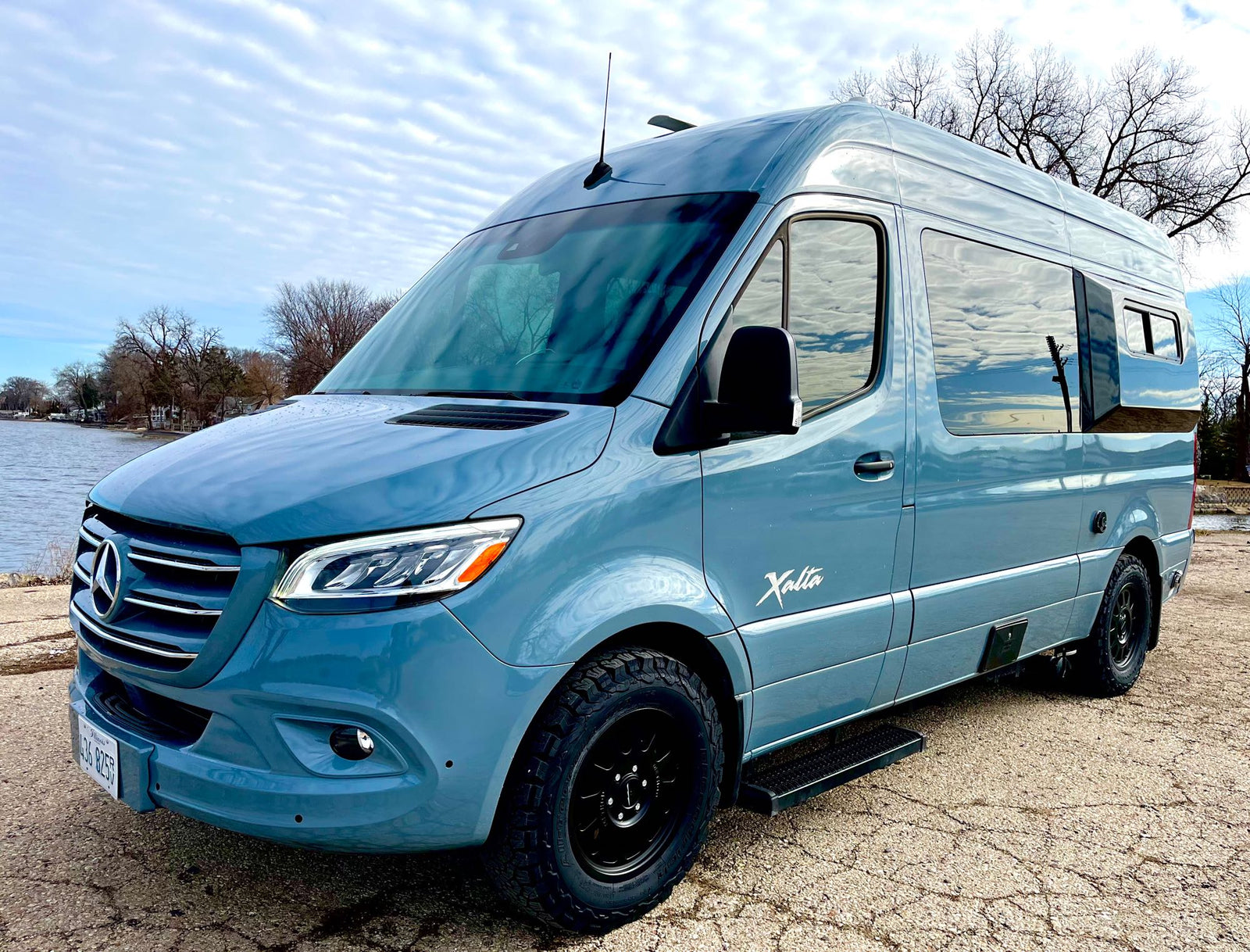 open doors to a serene lake view and blue sky. Perfect for van life adventures.|Regency RV rental: luxurious white quilted seats facing a beautiful ocean and beach view.|Step into this cozy RV rental bedroom: features a comfortable bed