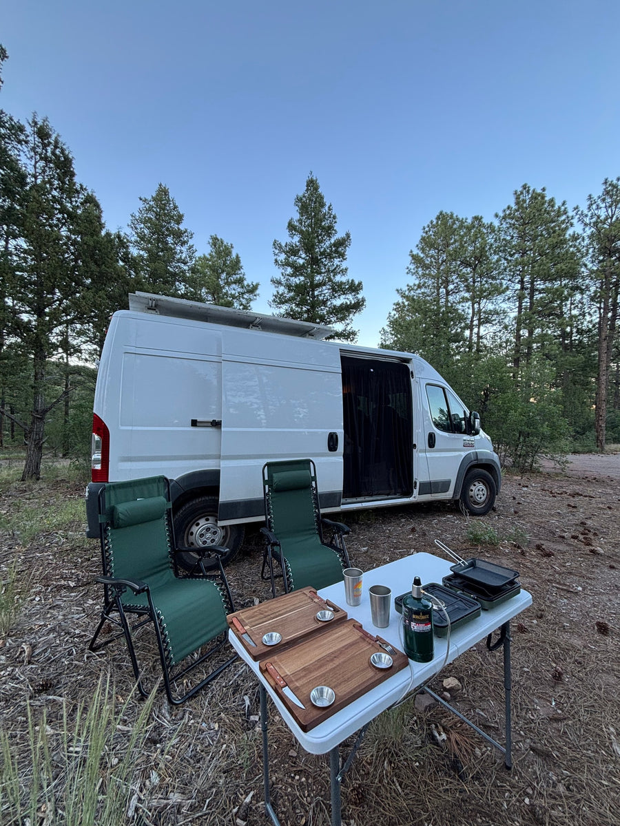 RV rental: Camper van in a forest, perfect for van life adventures, with chairs and outdoor cooking setup.
