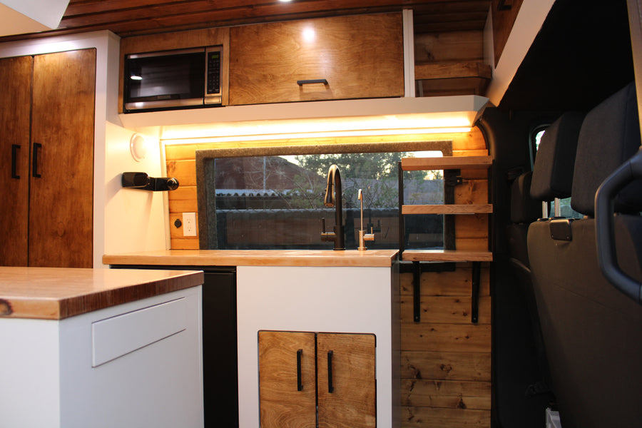 Cozy RV rental kitchen: wood cabinets, light counters, sink, microwave, and ambient lighting in a sleek van conversion.