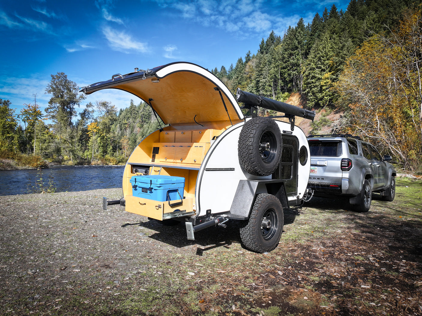 Experience outdoor adventure with this RV rental: a teardrop trailer with an open galley kitchen, hitched to an SUV by a scenic river.