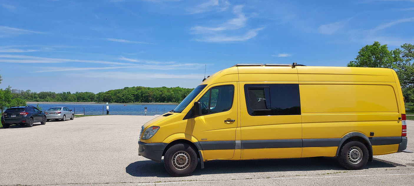 This vibrant yellow RV rental van is perfectly parked by a tranquil lake, surrounded by lush trees under a clear blue sky.