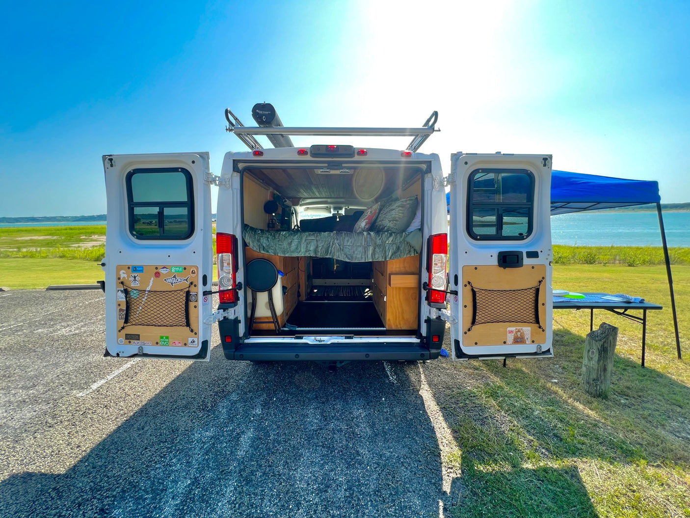 This rv rental camper van's open rear reveals custom interior, bed, storage, & roof shower, by a scenic lake. Ideal for van life.