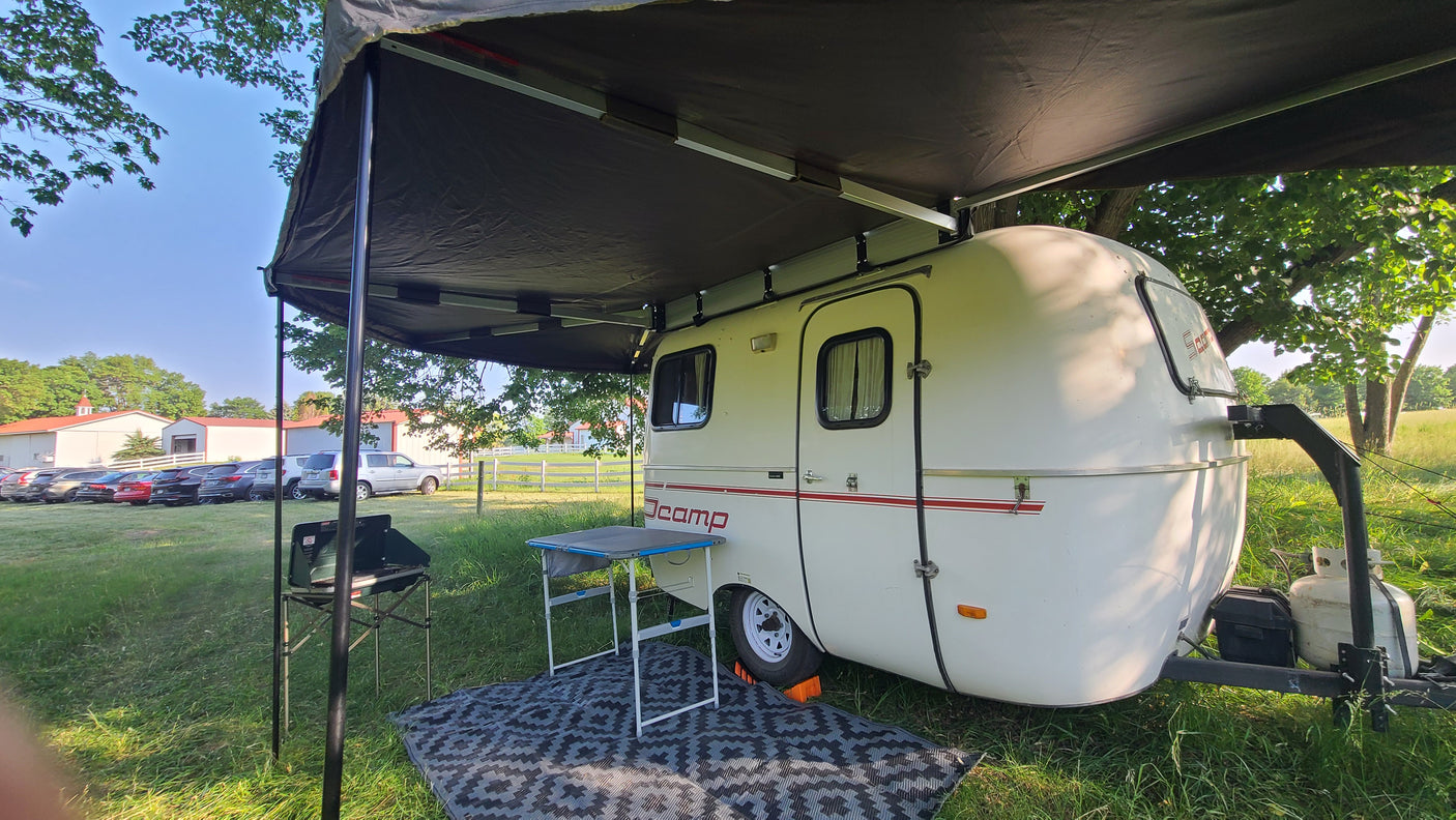 This white Jcamp RV rental camping trailer features an extended awning, outdoor table, and stove, set up on grass.