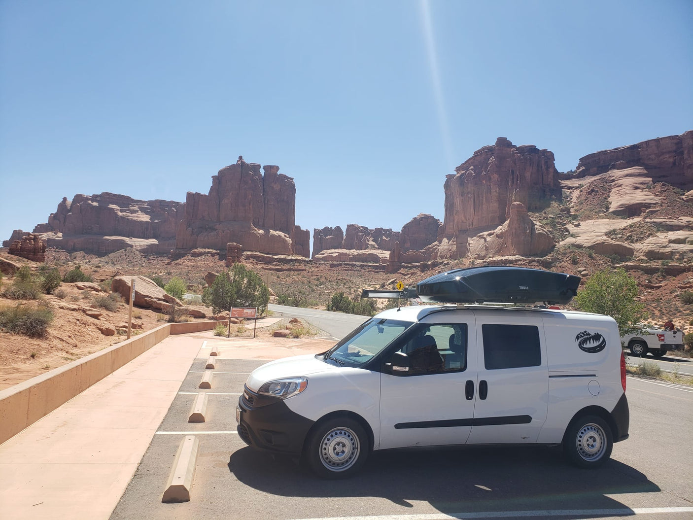 White van **RV rental** with rooftop storage box in a sunny desert parking area, backed by towering red rock formations.