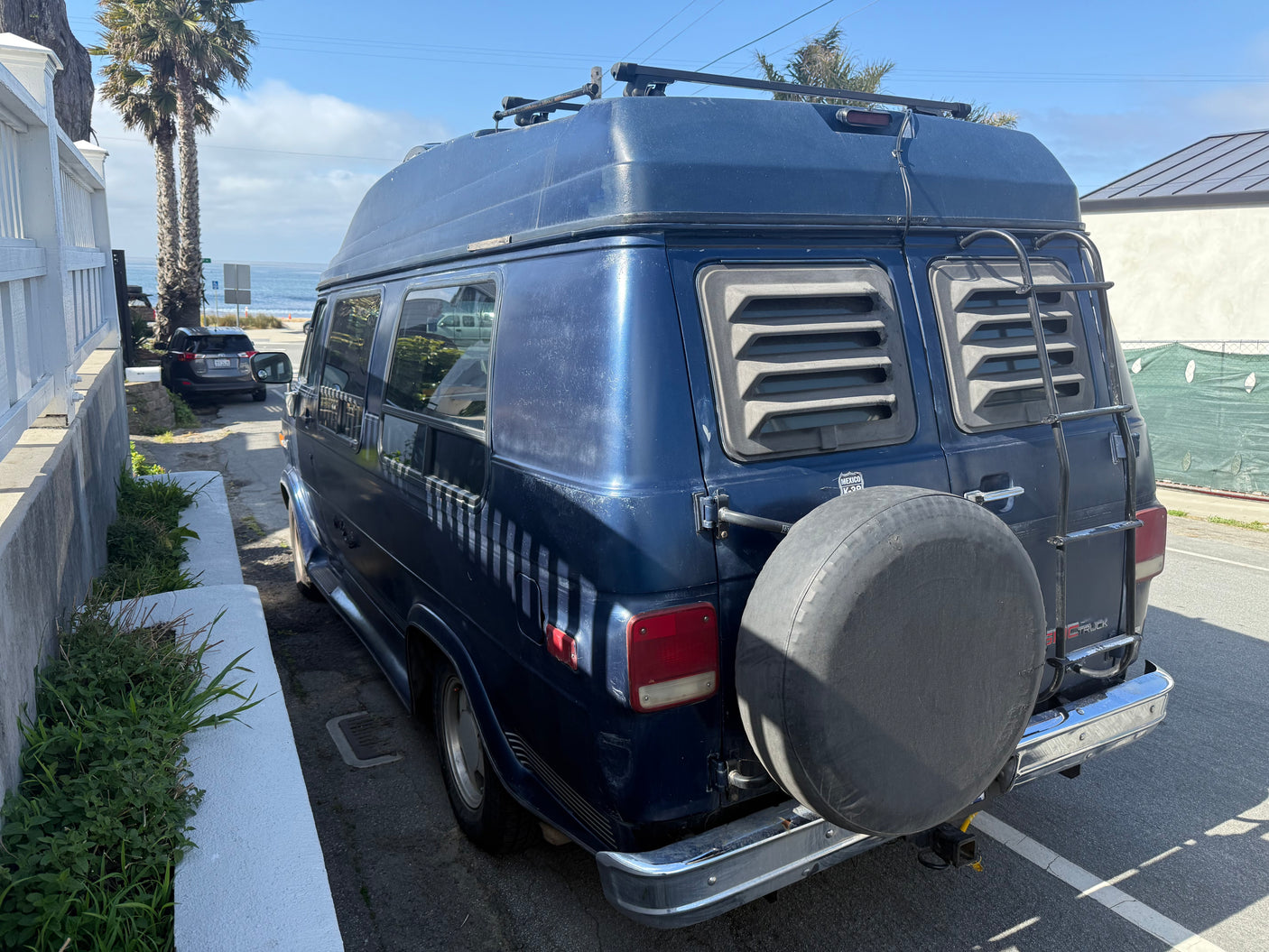 Blue RV rental van with a high top, rear ladder, and roof rack. Parked on a street overlooking the sunny ocean.