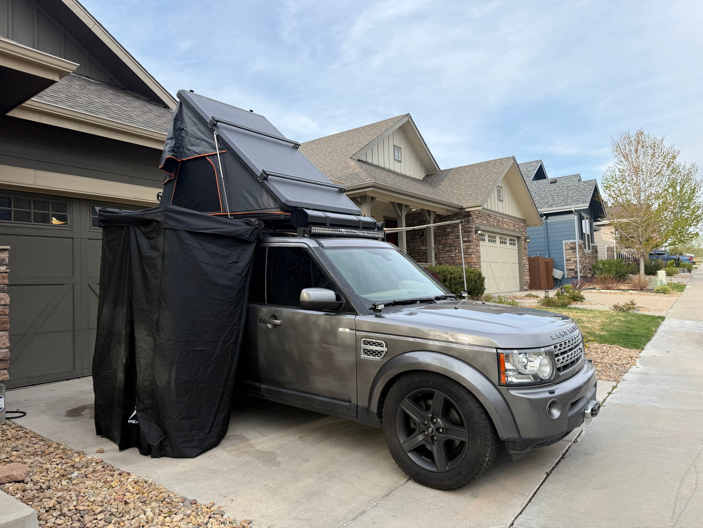 Grey Land Rover Discovery with an open rooftop tent and attached side privacy tent, parked in a driveway for an RV rental trip.