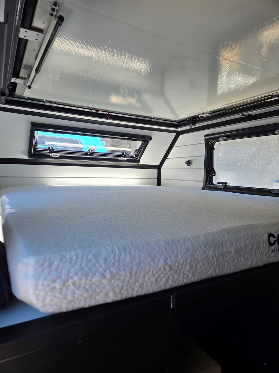 Compact RV rental sleeping area with textured mattress, large windows & pop-top roof, ready for adventure.