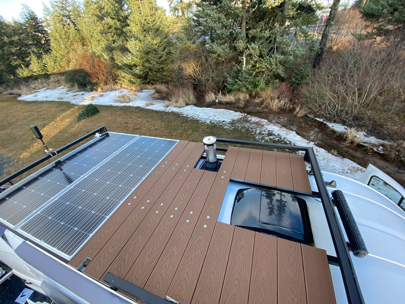 Rooftop of an rv rental with solar panels, a wooden deck, and a chimney pipe. Snow-dusted ground and trees are visible.