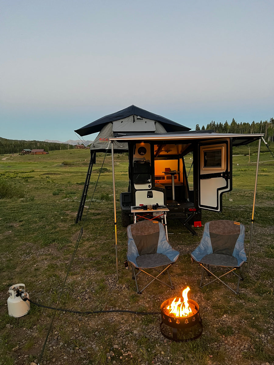 Twilight RV rental camping scene: a camper trailer with a rooftop tent, chairs, and a propane fire pit.