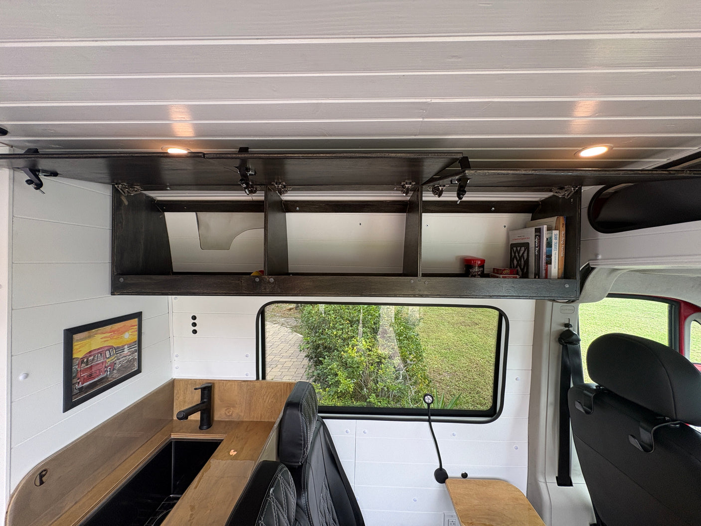 Cozy rv rental camper van interior: wood-slat ceiling, dark overhead storage with books, kitchenette, and a scenic window view.