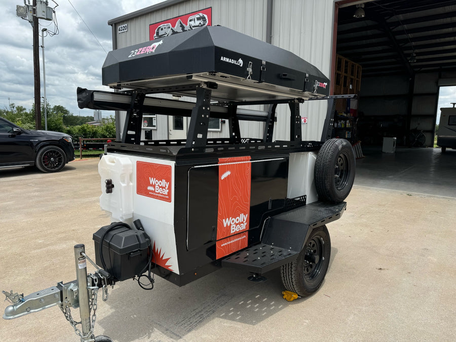 A sleek black and white Woolly Bear off-road utility trailer, topped with a 23ZERO rooftop tent, is shown, ideal for rv rental.