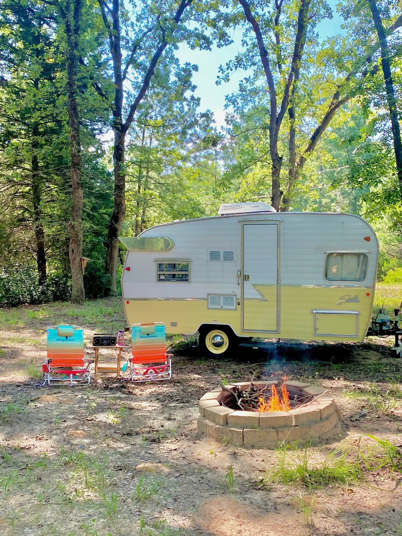 A cozy RV rental campsite with a yellow and white vintage camper, outdoor chairs, and a warm fire pit for glamping.