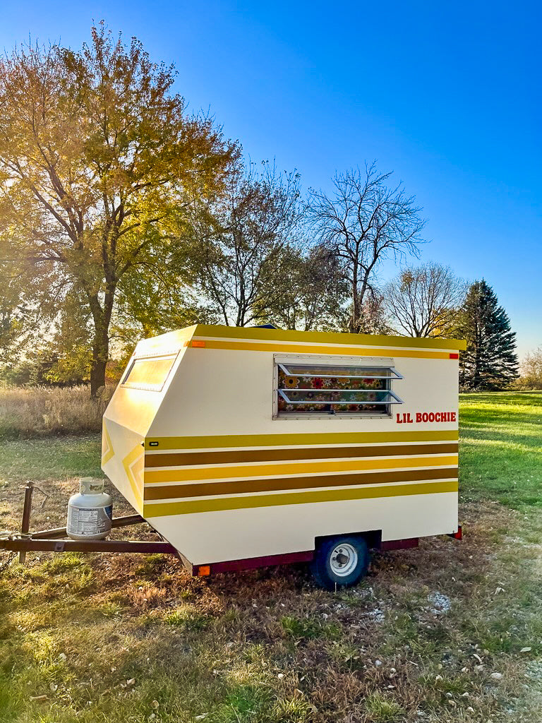 This cream & striped 'LIL BOOCHIE' vintage mini camper is an ideal rv rental, seen outdoors with autumn trees. Perfect for cozy travel.
