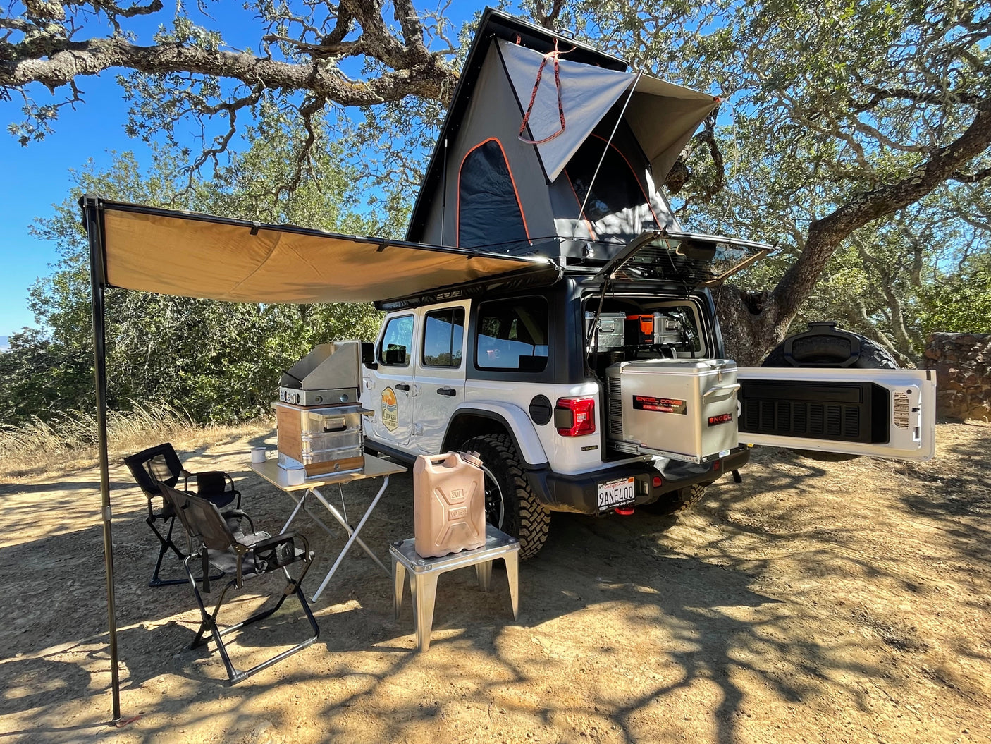 White Jeep RV rental fully equipped with rooftop tent, extended awning, outdoor kitchen, chairs, and fridge setup.