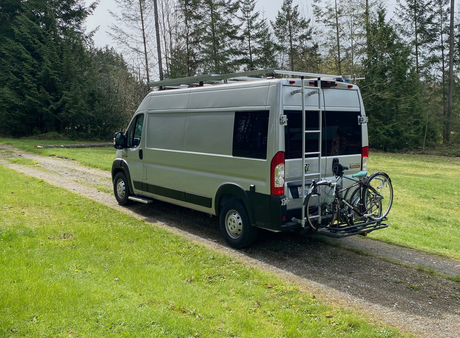 RV rental camper van with roof rack and bikes on a path. Tall trees & green grass for outdoor adventure.