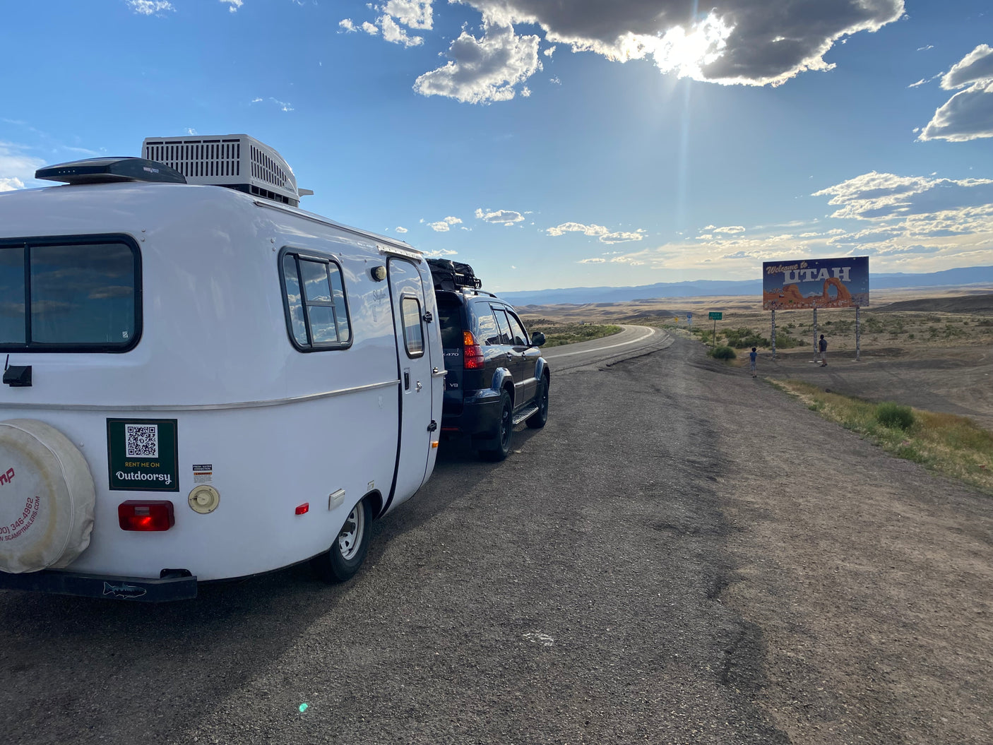 RV rental: White camper trailer & SUV on a Utah roadside; "Welcome to Utah" sign and scenic mountains.