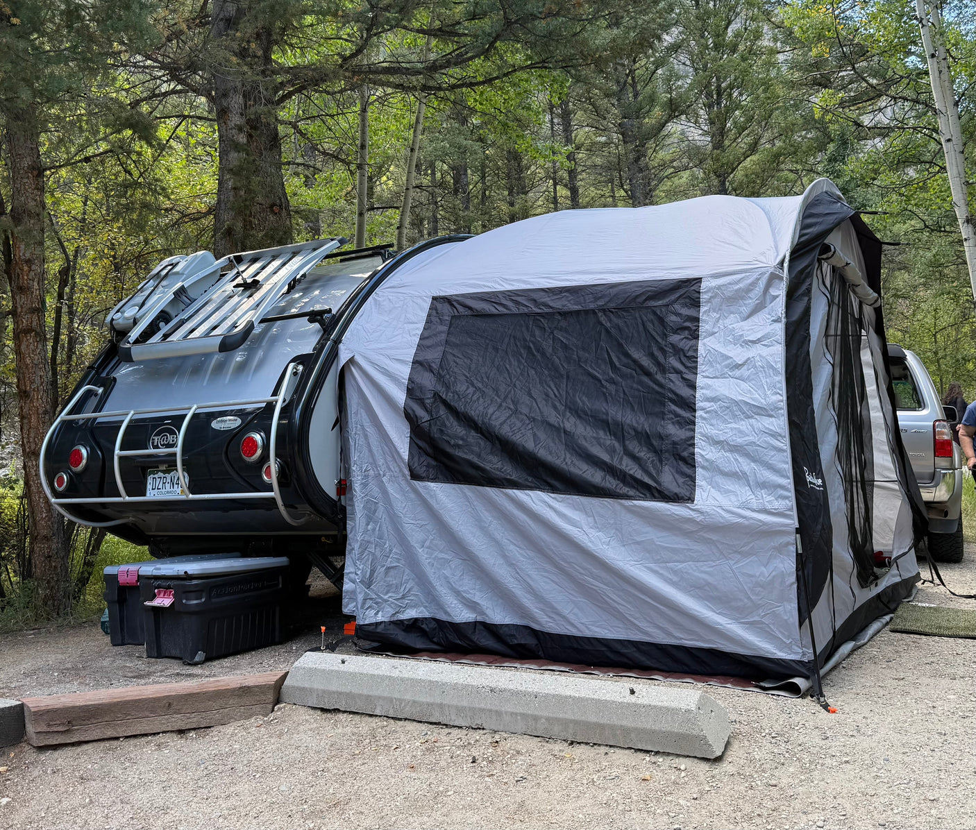 RV rental setup: A modern teardrop camper with an attached tent extension at a wooded campsite, ideal for outdoor adventures.