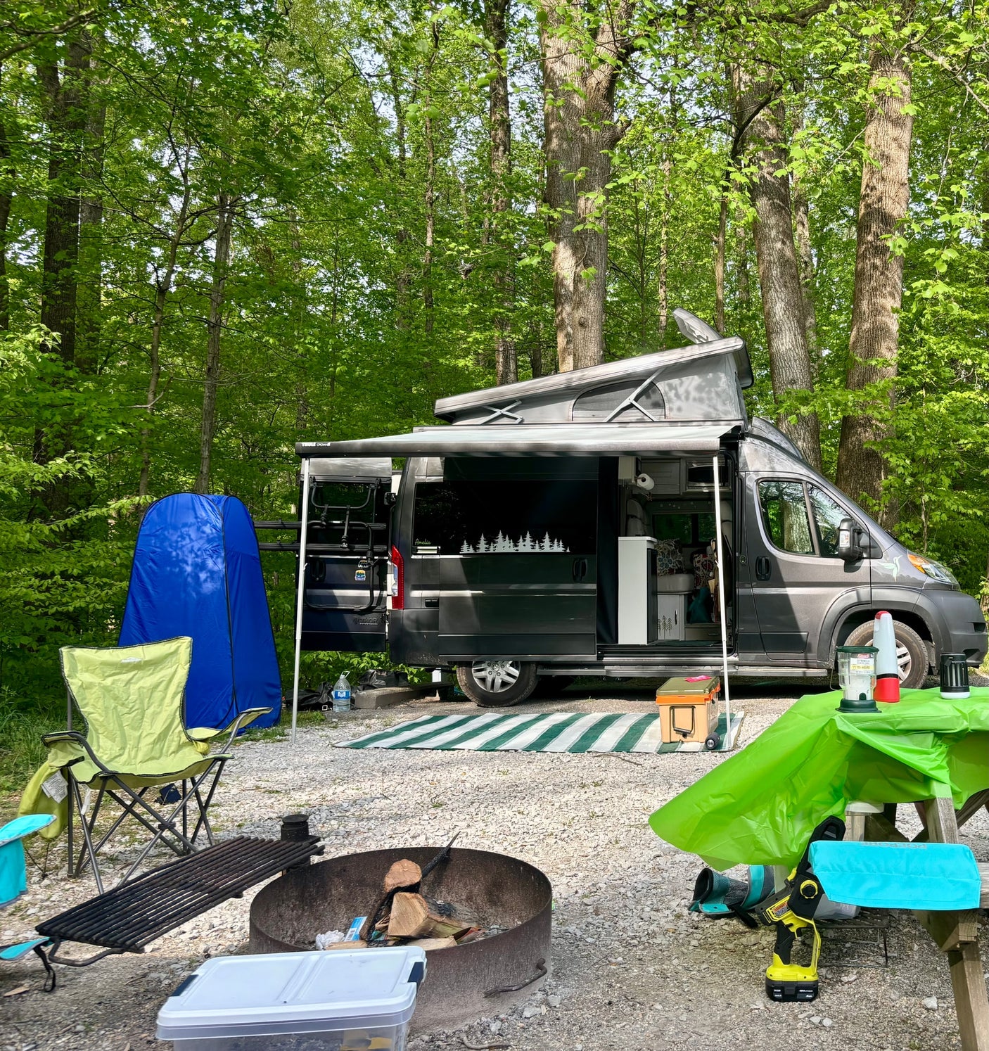 Serene forest camp setup featuring an RV rental with pop-top, awning, fire pit, and picnic area.