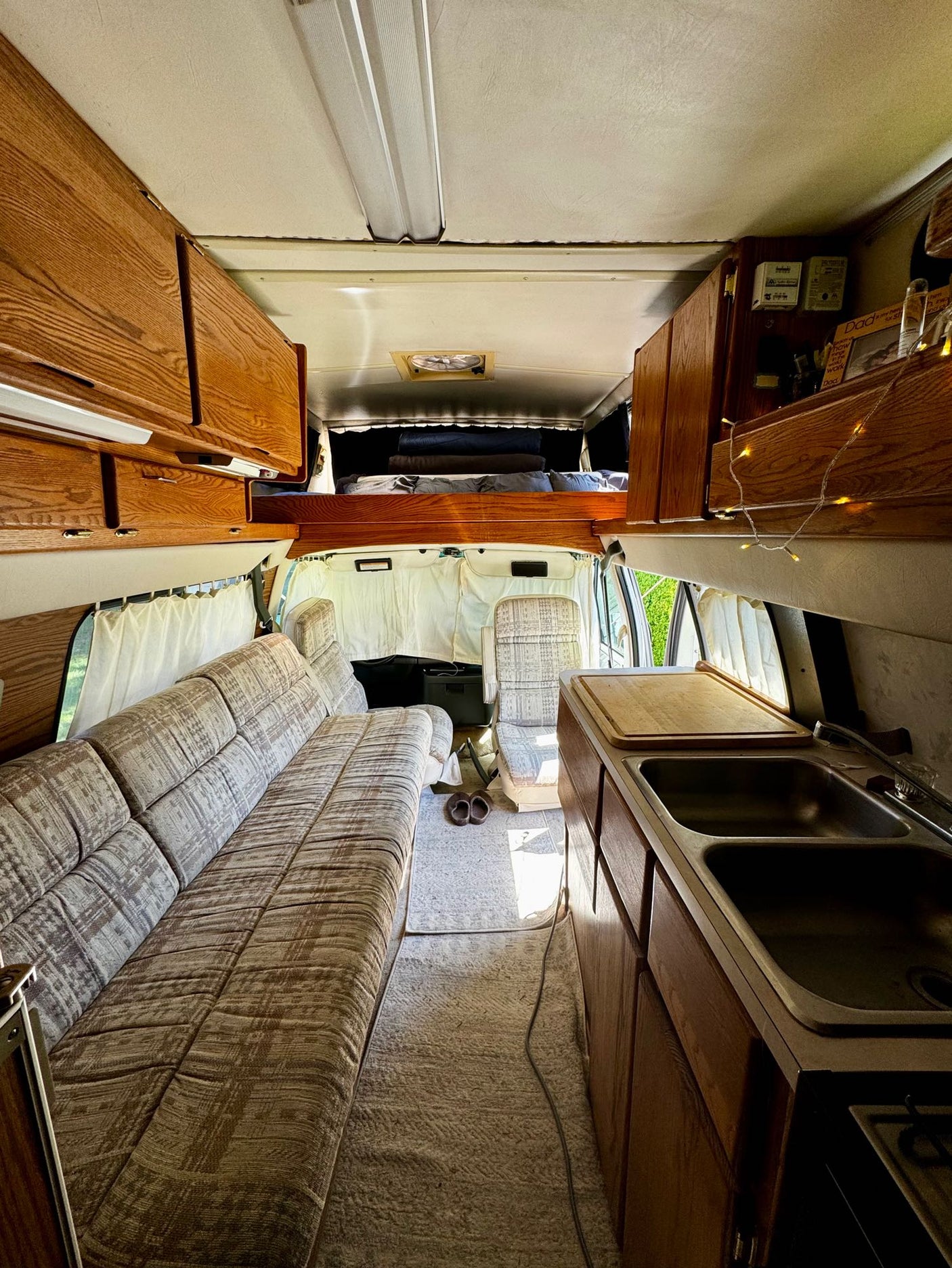 Inside this RV rental: a comfy sofa, kitchen with double sink, and bed. Wooden cabinets & light curtains create a cozy mobile home.
