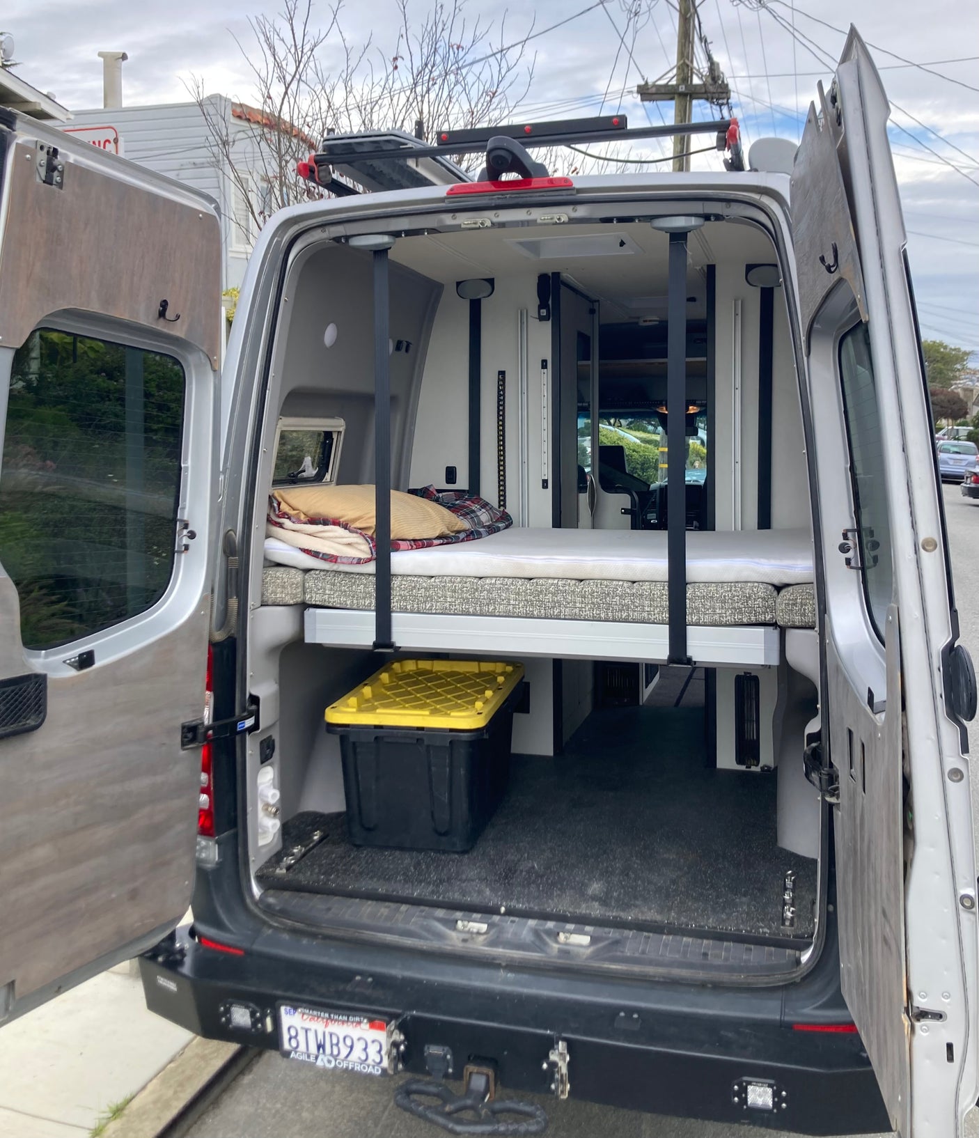 An RV rental van's open rear reveals a full bed setup, storage bin, and custom interior for road trips.