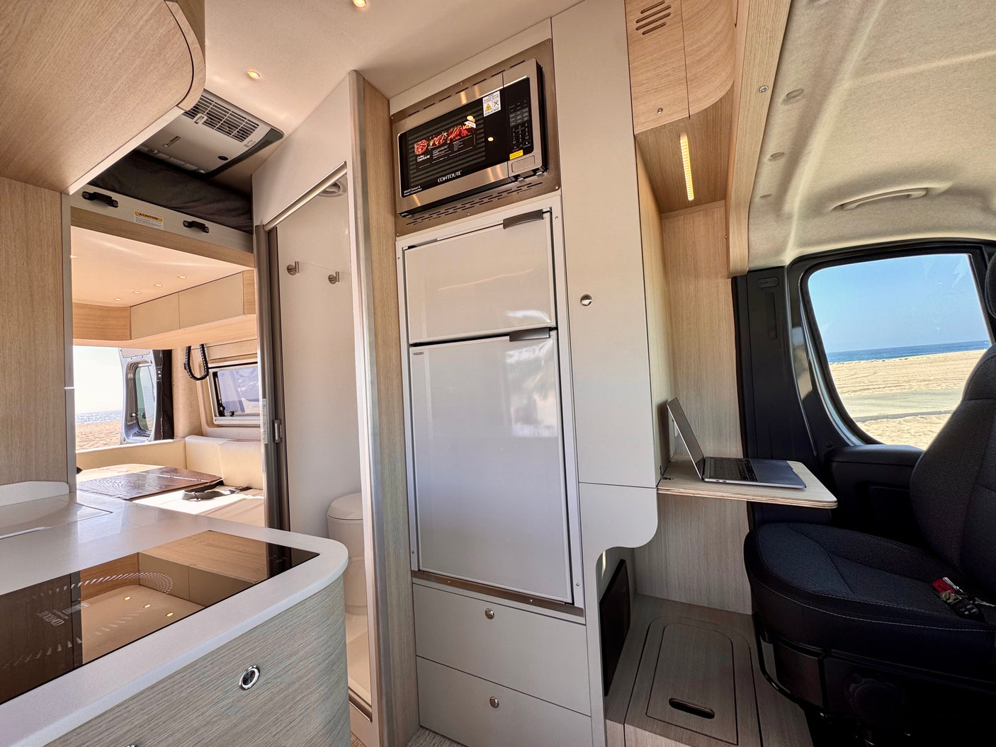 Rv rental interior: modern kitchen with microwave, fridge, and cooktop. Laptop desk by the window offers beach views.