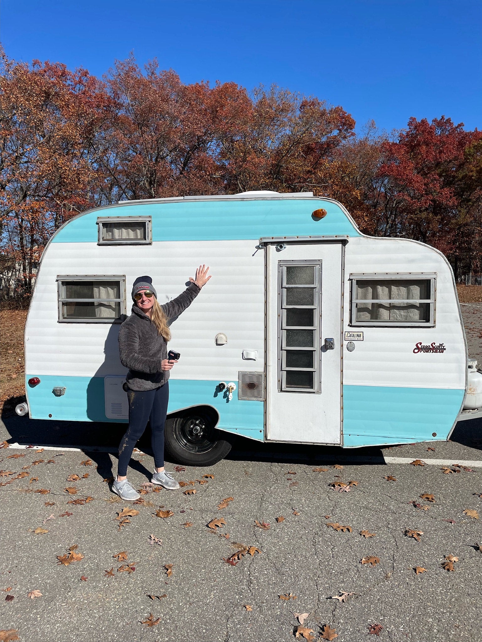 Happy woman waves by a vintage light blue & white Serro Scott Sportsman camper, ideal for an rv rental in autumn.