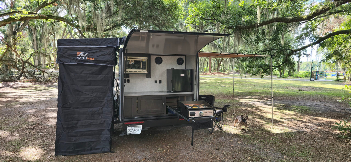 Compact RV rental with an open outdoor kitchen, microwave, fridge, and a shower tent. A dog enjoys the shady, mossy campsite.