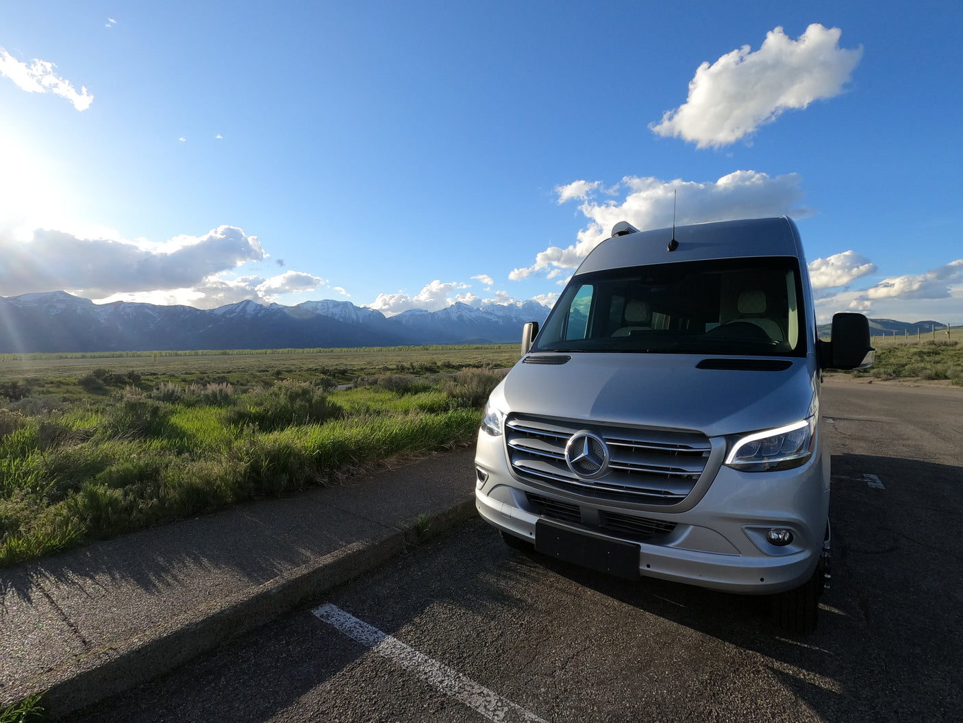 Silver Mercedes Sprinter RV rental parked in a stunning landscape with snowy mountains & green fields.