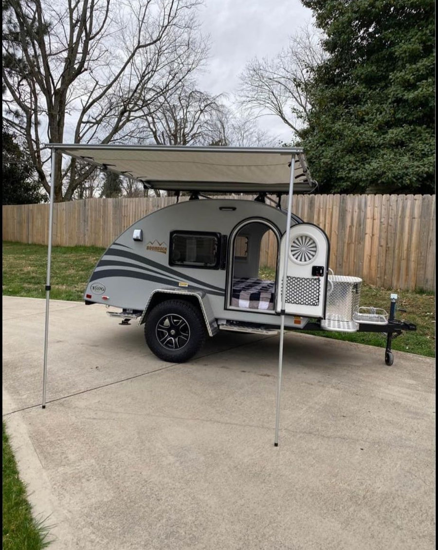 Exterior view of a grey teardrop RV rental with an extended awning. The open door reveals a cozy checkered bed inside.