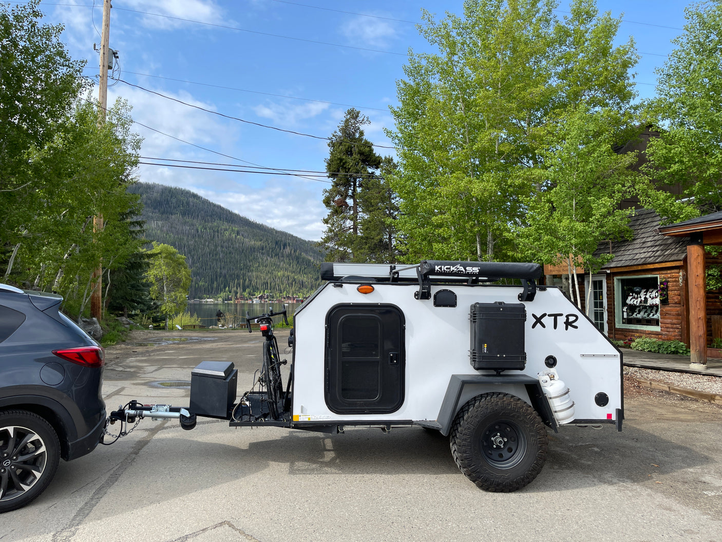 White XTR off-road camper hitched to a grey SUV near a lake & mountains. Perfect for your next rv rental adventure.