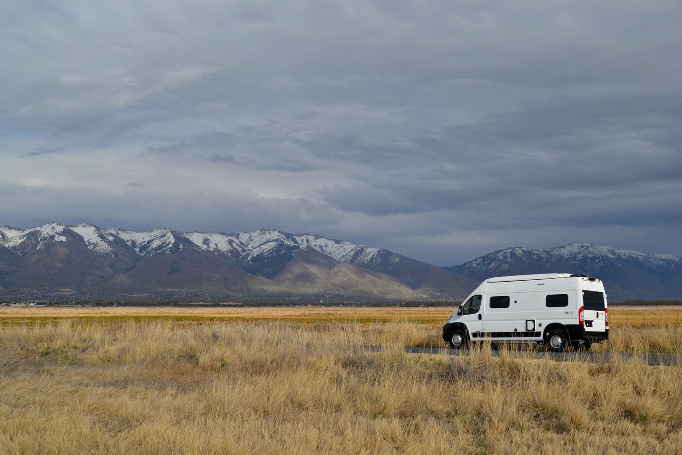 This white rv rental rests amidst golden fields, framed by majestic snow-capped mountains under a dramatic sky.