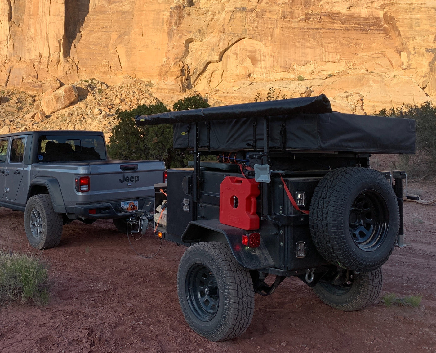 Gray Jeep Gladiator towing an off-road overland trailer with rooftop tent by desert cliffs. Perfect for adventure **RV rental**.