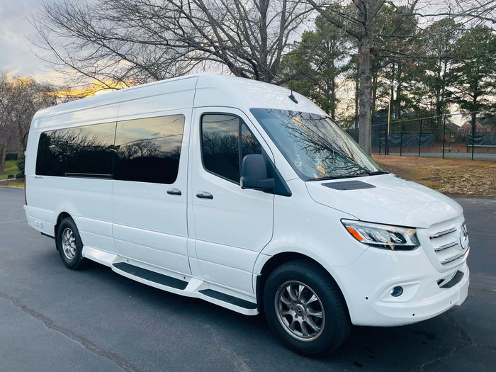 is parked on a gravel lot with trees and old structures behind it.|White Mercedes Sprinter van. A spacious rv rental parked among sunlit trees