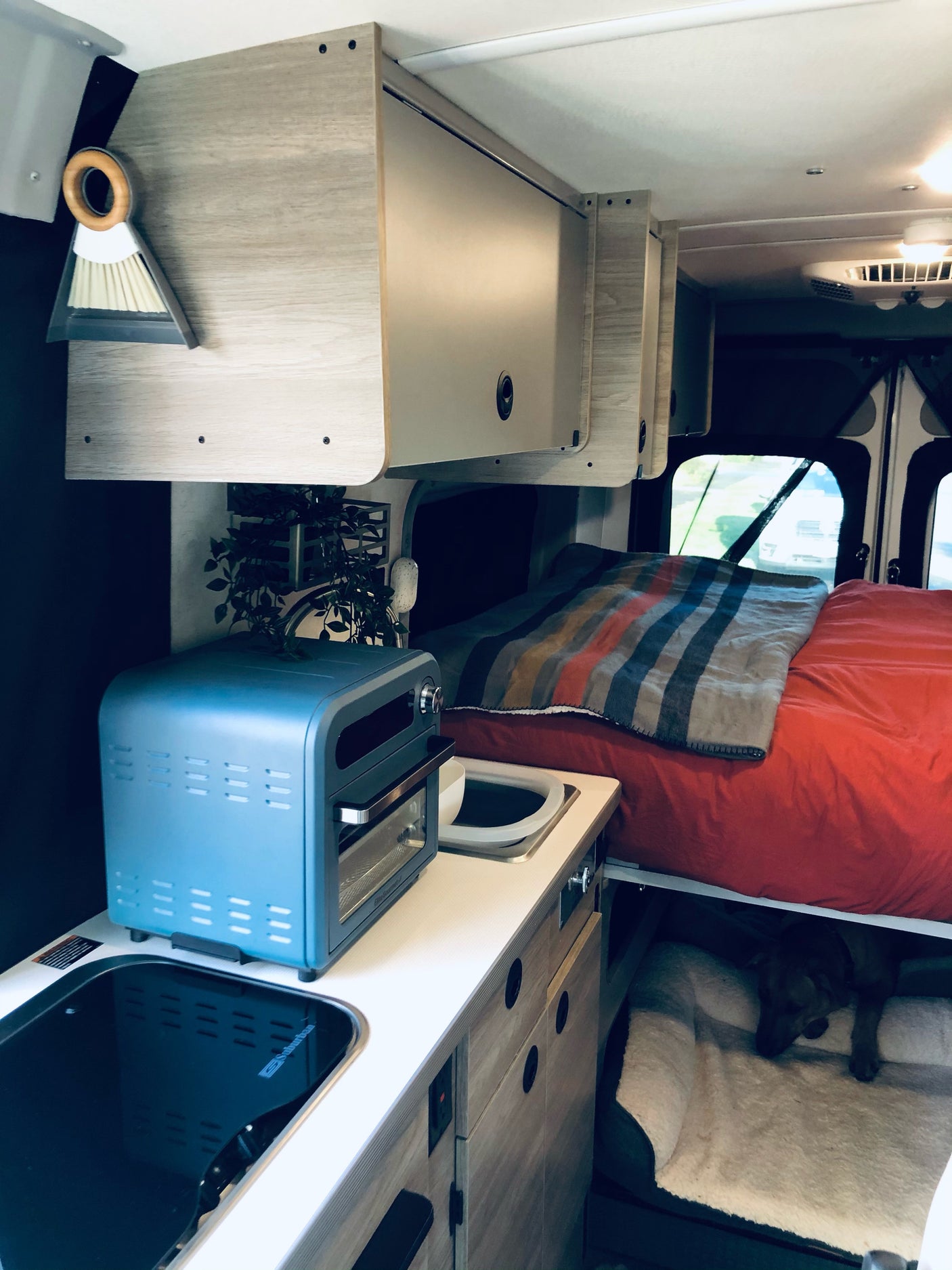 This cozy RV rental interior features a compact kitchenette, bed with red linens, overhead storage, and a dog's bed below.